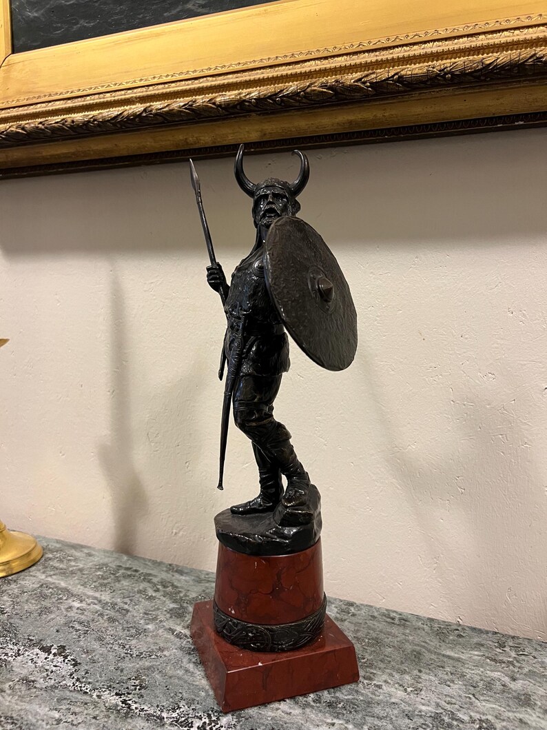 May include: A bronze sculpture of a Viking warrior holding a spear and shield. The sculpture is mounted on a red marble base.