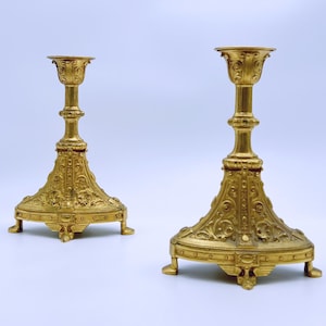 May include: Two ornate, gold-colored candle holders. Each has a detailed base with a flared design and three small feet. The central column is intricately carved, leading to a cup-shaped top. The candle holders are likely made of metal.