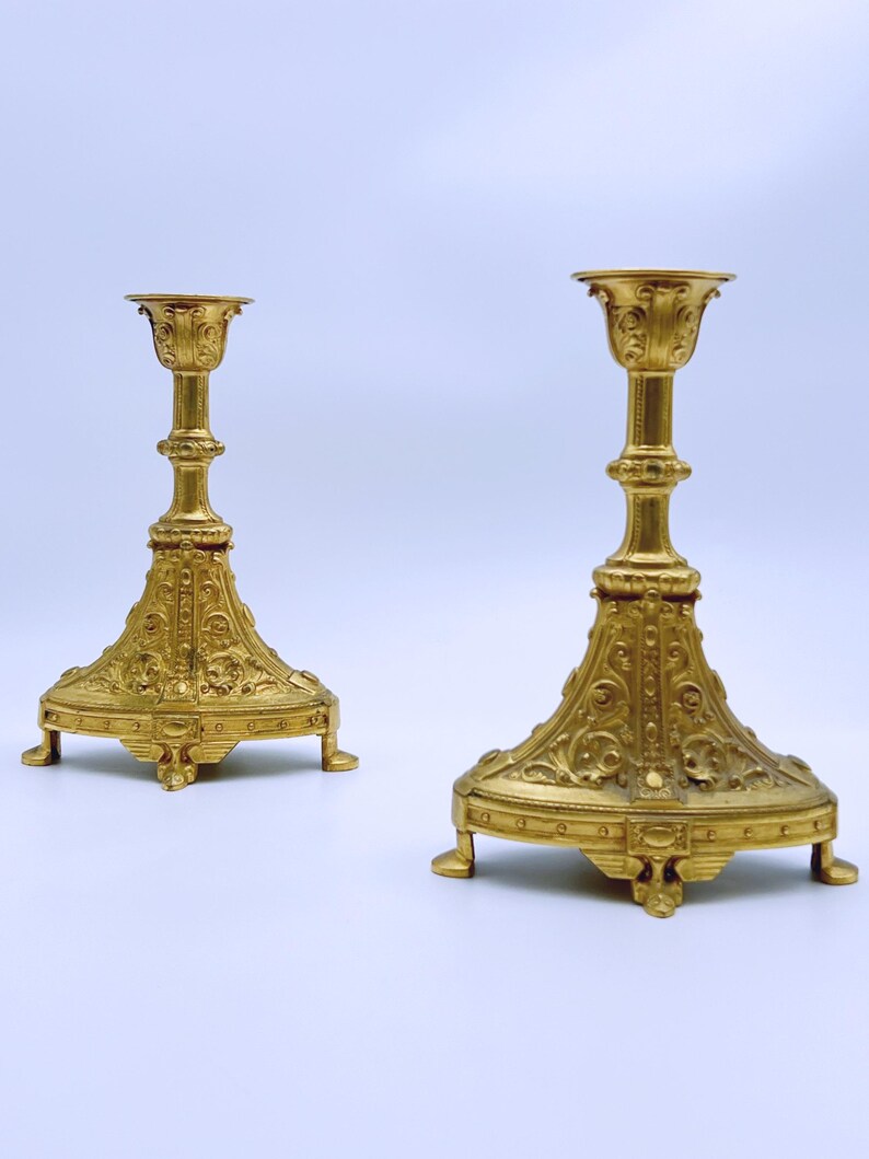 May include: Two ornate, gold-colored candle holders. Each has a detailed base with a flared design and three small feet. The central column is intricately carved, leading to a cup-shaped top. The candle holders are likely made of metal.