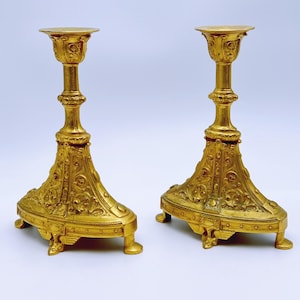 May include: Two ornate, gold-colored candle holders. Each has a wide, decorative base with intricate patterns and a tall, slender stem leading to a circular top. The candle holders are symmetrical and stand on a white background.