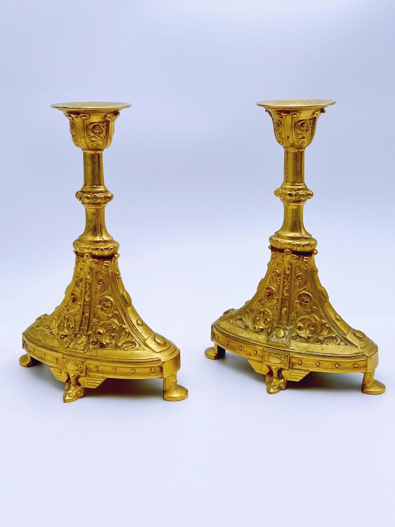 May include: Two ornate, gold-colored candle holders. Each has a wide, decorative base with intricate patterns and a tall, slender stem leading to a circular top. The candle holders are symmetrical and stand on a white background.
