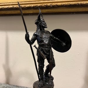 May include: A bronze sculpture of a warrior holding a spear and shield. The warrior is wearing a helmet with horns and a tunic. The sculpture is mounted on a red marble base.