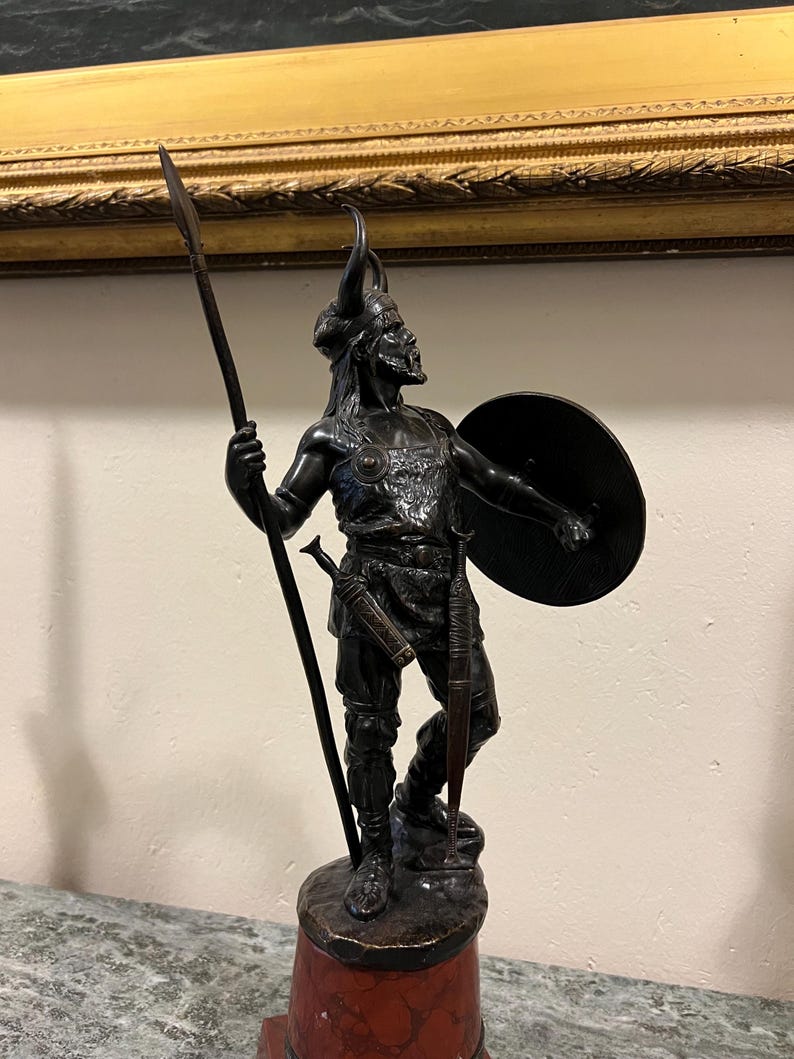 May include: A bronze sculpture of a warrior holding a spear and shield. The warrior is wearing a helmet with horns and a tunic. The sculpture is mounted on a red marble base.