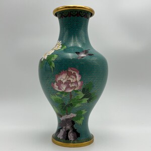 Puede incluir: Un jarr&oacute;n decorativo de color turquesa con borde y base dorados. El jarr&oacute;n presenta un dise&ntilde;o floral con una gran peon&iacute;a rosa, flores blancas y un peque&ntilde;o p&aacute;jaro. El jarr&oacute;n tiene una superficie texturizada.