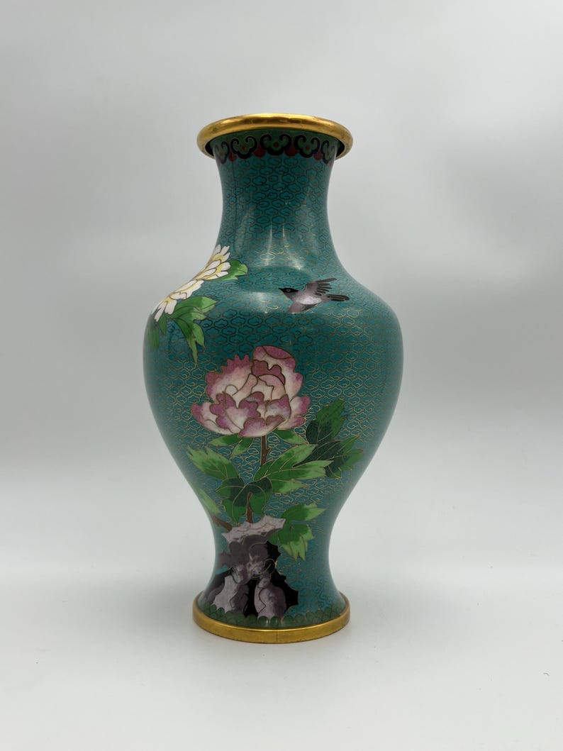 Puede incluir: Un jarr&oacute;n decorativo de color turquesa con borde y base dorados. El jarr&oacute;n presenta un dise&ntilde;o floral con una gran peon&iacute;a rosa, flores blancas y un peque&ntilde;o p&aacute;jaro. El jarr&oacute;n tiene una superficie texturizada.
