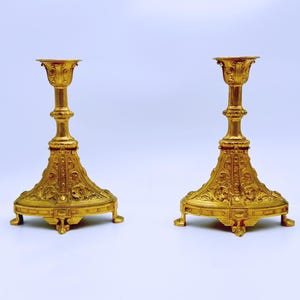 May include: Two ornate gold-colored candlesticks. Each candlestick has a wide, decorative base with intricate patterns and a tall, slender stem leading to a cup-shaped top. The candlesticks are symmetrical and stand on a white background.