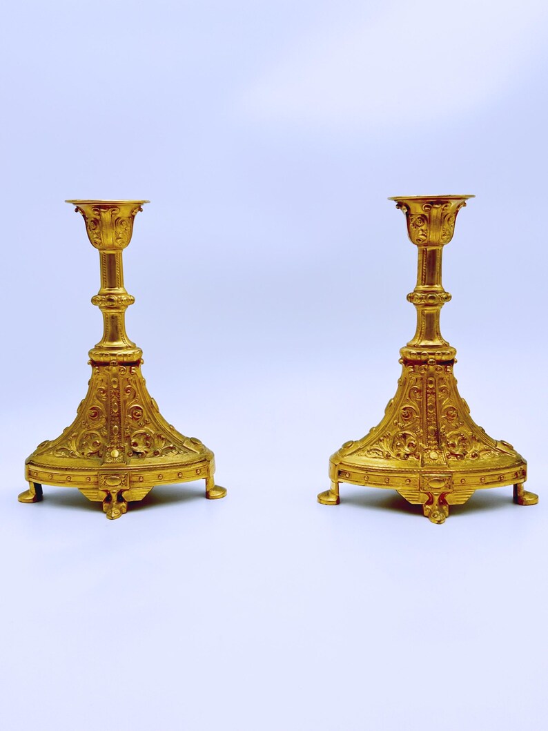 May include: Two ornate gold-colored candlesticks. Each candlestick has a wide, decorative base with intricate patterns and a tall, slender stem leading to a cup-shaped top. The candlesticks are symmetrical and stand on a white background.