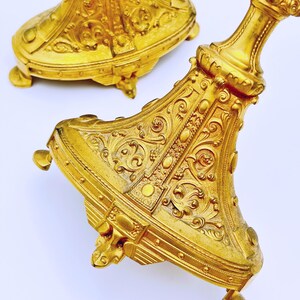May include: Two ornate, gold-colored candle holders with intricate embossed designs. The bases have a detailed, floral-like pattern, and the stems are embellished with decorative elements. The candle holders are on a white background.