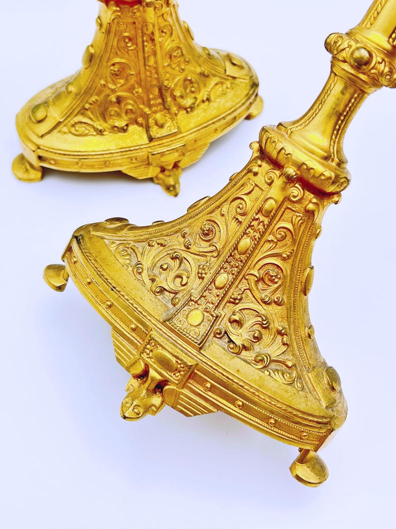 May include: Two ornate, gold-colored candle holders with intricate embossed designs. The bases have a detailed, floral-like pattern, and the stems are embellished with decorative elements. The candle holders are on a white background.