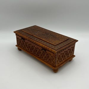 May include: A rectangular wooden box with intricate carved floral and geometric patterns. The box is a warm brown color, with a hinged lid and small feet. The carvings are detailed and create a textured surface.