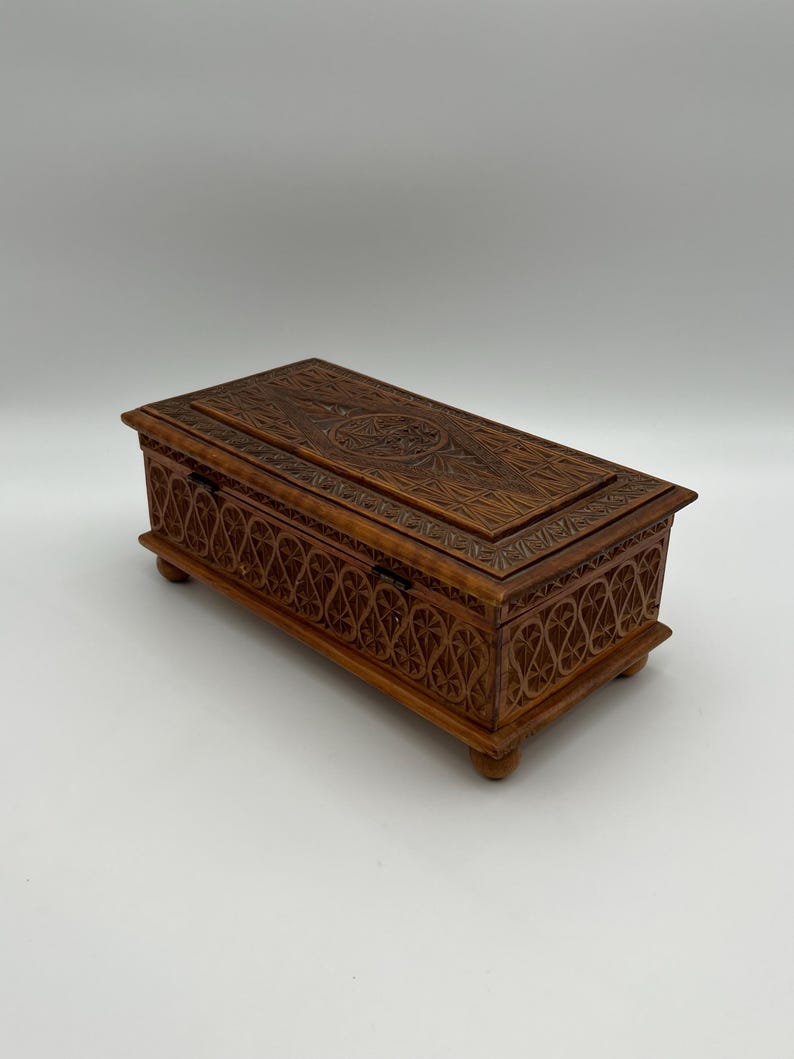 May include: A rectangular wooden box with intricate carved floral and geometric patterns. The box is a warm brown color, with a hinged lid and small feet. The carvings are detailed and create a textured surface.