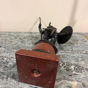 May include: A black metal statue of a warrior holding a spear and shield, standing on a red marble base.