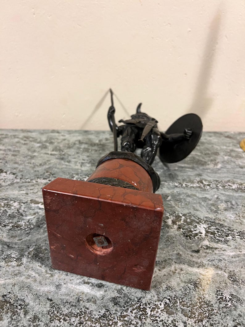 May include: A black metal statue of a warrior holding a spear and shield, standing on a red marble base.