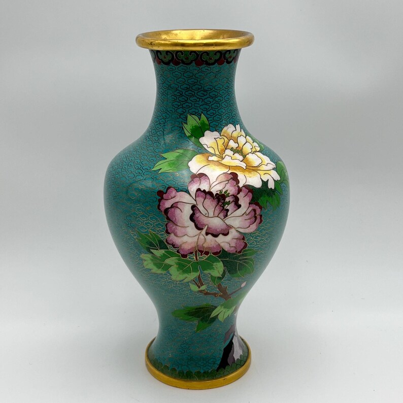 Puede incluir: Un jarr&oacute;n de cloisonn&eacute; turquesa con borde y base dorados. El jarr&oacute;n presenta un dise&ntilde;o floral con peon&iacute;as rosas y amarillas y hojas verdes. El jarr&oacute;n tiene una superficie texturizada.