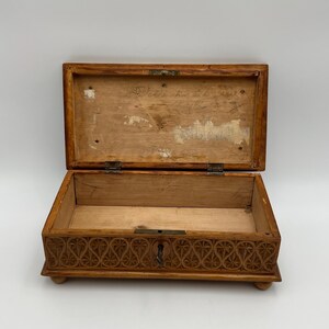 May include: Open wooden box with a hinged lid. The box is light brown with a carved floral design on the front. The interior is plain wood. The lid has text written on it. The box is resting on small, rounded feet.