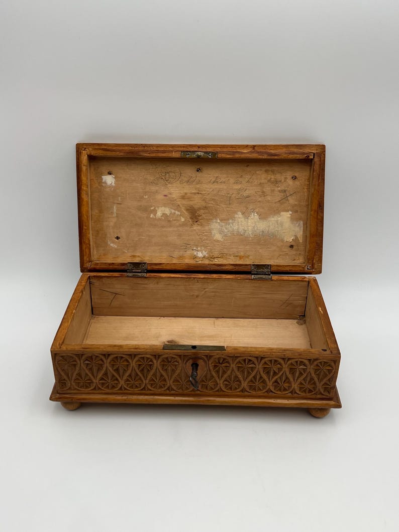 May include: Open wooden box with a hinged lid. The box is light brown with a carved floral design on the front. The interior is plain wood. The lid has text written on it. The box is resting on small, rounded feet.