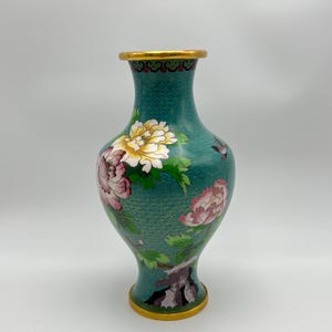 Puede incluir: Un jarr&oacute;n de cloisonn&eacute; turquesa con borde y base dorados. El jarr&oacute;n presenta un dise&ntilde;o floral con peon&iacute;as rosas y blancas y hojas verdes. El jarr&oacute;n tiene una superficie texturizada.