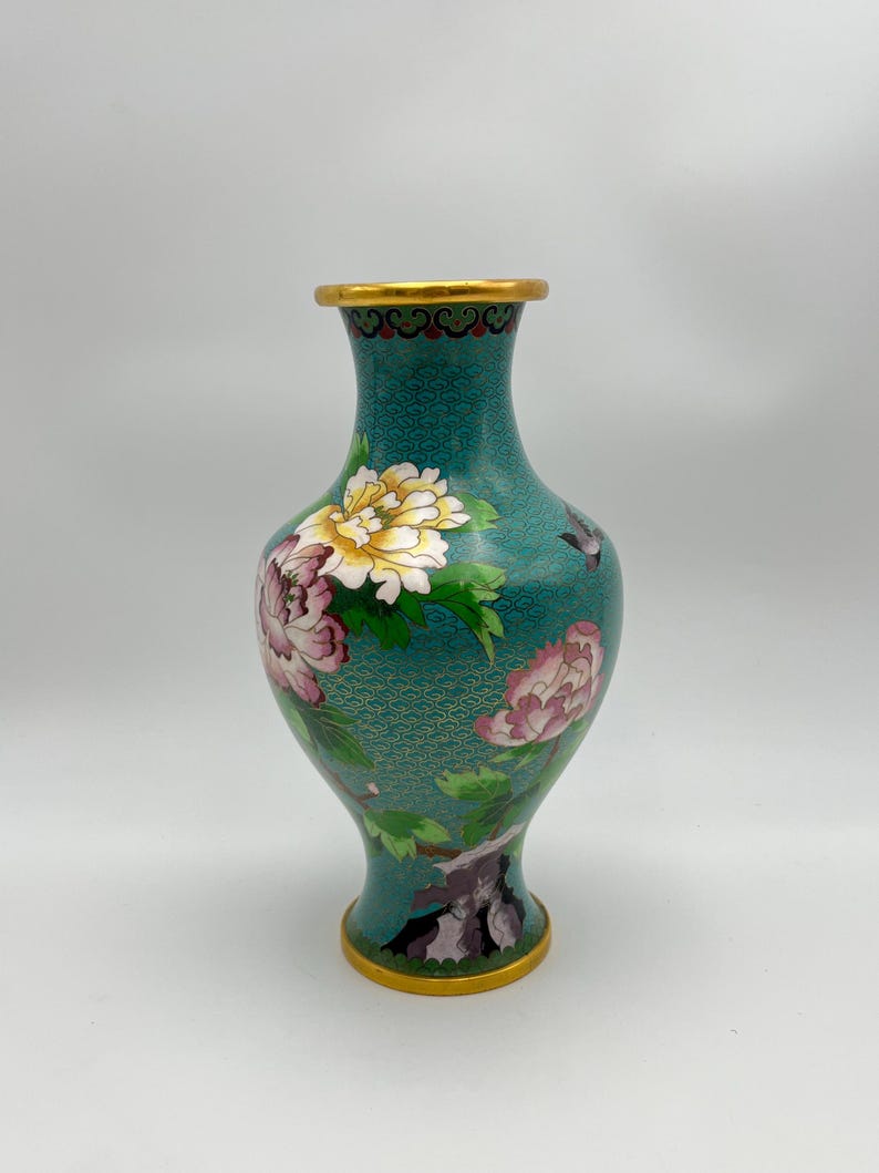 Puede incluir: Un jarr&oacute;n de cloisonn&eacute; turquesa con borde y base dorados. El jarr&oacute;n presenta un dise&ntilde;o floral con peon&iacute;as rosas y blancas y hojas verdes. El jarr&oacute;n tiene una superficie texturizada.