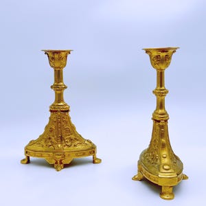 May include: Two ornate, gold-colored candle holders. Each has a detailed base with a decorative pattern, a slender stem, and a cup-shaped top. The candle holders are made of metal and have a shiny finish.
