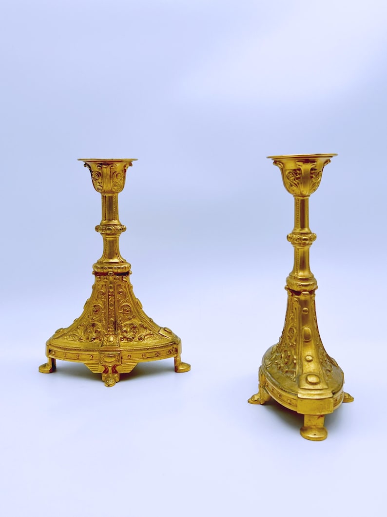 May include: Two ornate, gold-colored candle holders. Each has a detailed base with a decorative pattern, a slender stem, and a cup-shaped top. The candle holders are made of metal and have a shiny finish.