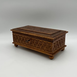 May include: A rectangular wooden box with intricate carved floral and geometric patterns. The box is a warm brown color, with a small drawer and a decorative keyhole. It rests on four small, rounded feet.