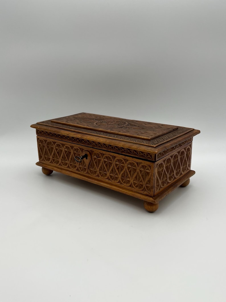 May include: A rectangular wooden box with intricate carved floral and geometric patterns. The box is a warm brown color, with a small drawer and a decorative keyhole. It rests on four small, rounded feet.
