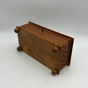 May include: A rectangular wooden box with intricate carved floral patterns. The box has a hinged lid and rests on four rounded feet. A small metal latch is visible on the top. The wood has a warm, natural tone.