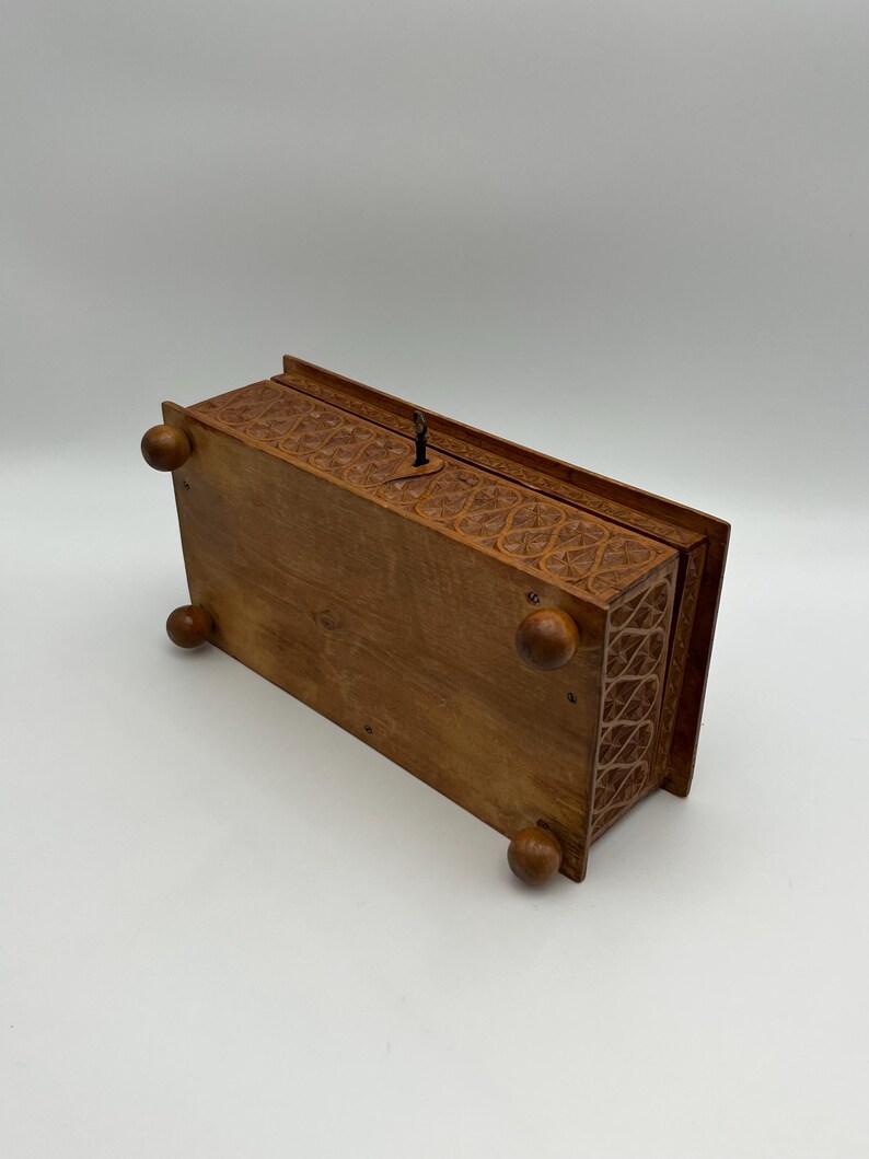May include: A rectangular wooden box with intricate carved floral patterns. The box has a hinged lid and rests on four rounded feet. A small metal latch is visible on the top. The wood has a warm, natural tone.