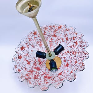 May include: A white glass ceiling light fixture with a red speckled pattern and a ruffled edge. The fixture has three light bulbs and a gold metal hanging mechanism.