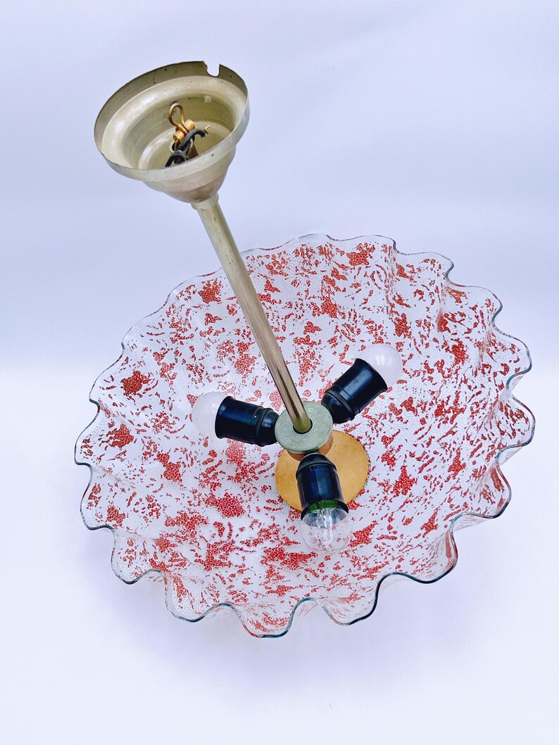 May include: A white glass ceiling light fixture with a red speckled pattern and a ruffled edge. The fixture has three light bulbs and a gold metal hanging mechanism.