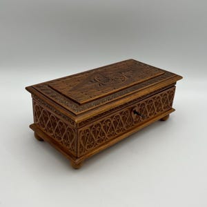 May include: A rectangular wooden box with intricate carved floral and geometric patterns. The box is a warm brown color, with a keyhole and small feet. The lid features a detailed carved design, and the sides have a repeating pattern.
