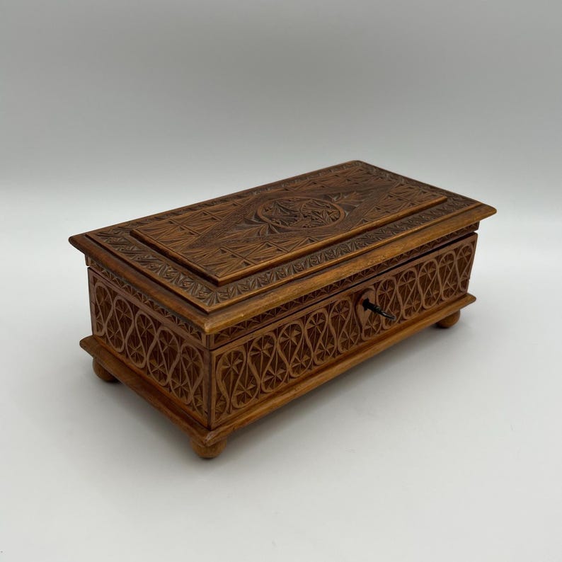 May include: A rectangular wooden box with intricate carved floral and geometric patterns. The box is a warm brown color, with a keyhole and small feet. The lid features a detailed carved design, and the sides have a repeating pattern.
