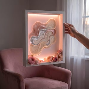 Peut inclure: Une œuvre d'art encadrée de blanc avec un motif abstrait dans des tons de rose, beige et argent. L'œuvre est éclairée de l'intérieur et ornée de roses roses à la base. Une main tient l'œuvre.