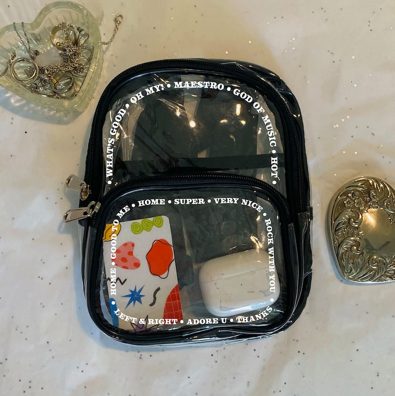 May include: A clear plastic backpack with black trim and white text around the edges. The text reads "What's good to me, Oh my! Maestro, God of Music, Hot", "Home, Super, Very Nice, Rock With You", and "Left & Right, Adore U, Thanks". The backpack is filled with various items, including a card with a colourful abstract design, a pair of white earbuds, and a black object.