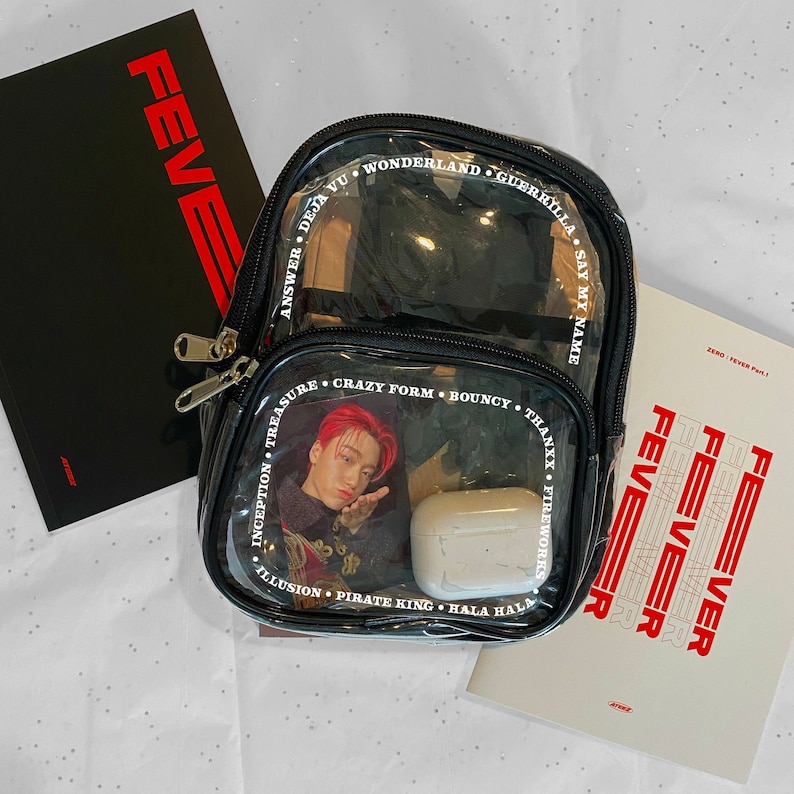 May include: A clear plastic backpack with black straps and a black zipper. The backpack has a white border with the names of songs printed around the outside. The backpack has a white case inside with a picture of a person with red hair. The text on the backpack reads "Answer", "Deja Vu", "Wonderland", "Guerrilla", "Say My Name", "Inception", "Treasure", "Crazy Form", "Bouncy", "Thanxx", "Fireworks", "Illusion", "Pirate King", "Hala Hala".