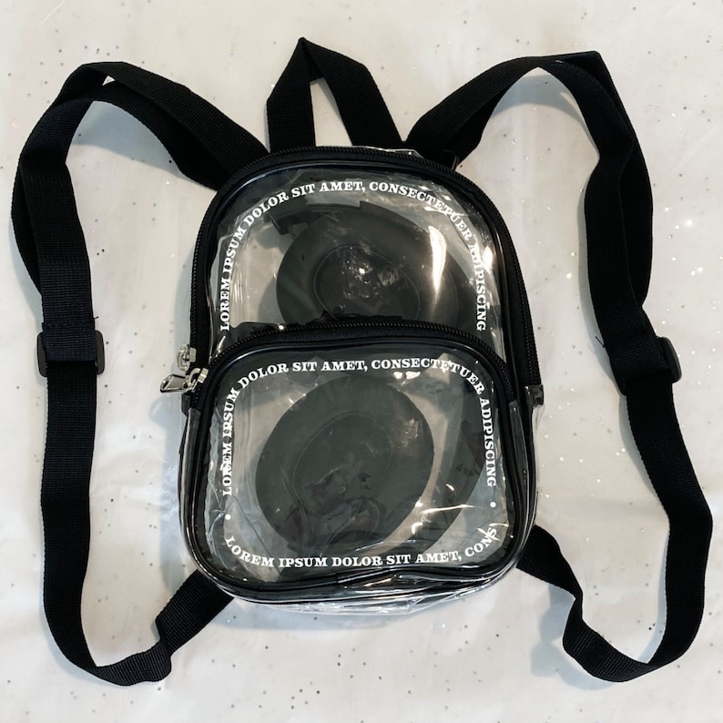 May include: A clear plastic backpack with black adjustable straps. The backpack has two clear compartments with black trim and white text that reads "LOREM IPSUM DOLOR SIT AMET, CONSECTETUER ADIPISCING".