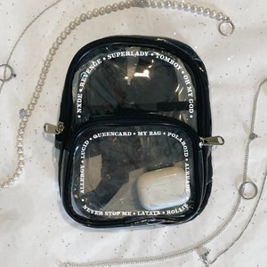 May include: A black clear plastic backpack with white text around the edges. The text reads "SUPERLADY TOMBOY OH MY GOD", "NXDEREVENGE", "QUEENCARD. MY BAG. POLAROID. ALREADY", "ALLERGY LUCID", "NEVER STOP ME LATATA. ROLLIE".