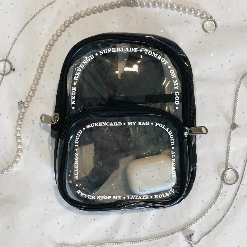 May include: A black clear plastic backpack with white text around the edges. The text reads "SUPERLADY TOMBOY OH MY GOD", "NXDEREVENGE", "QUEENCARD. MY BAG. POLAROID. ALREADY", "ALLERGY LUCID", "NEVER STOP ME LATATA. ROLLIE".