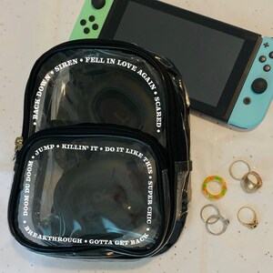 May include: A black clear plastic backpack with white text around the edges. The text reads "Back Down Siren Fell In Love Again Scared. Doom Du Doom Jump Killin' It Do It Like This Super Chic Breakthrough Gotta Get Back".