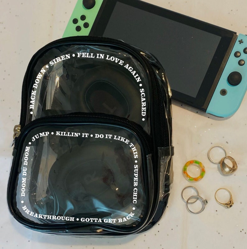 May include: A black clear plastic backpack with white text around the edges. The text reads "Back Down Siren Fell In Love Again Scared. Doom Du Doom Jump Killin' It Do It Like This Super Chic Breakthrough Gotta Get Back".