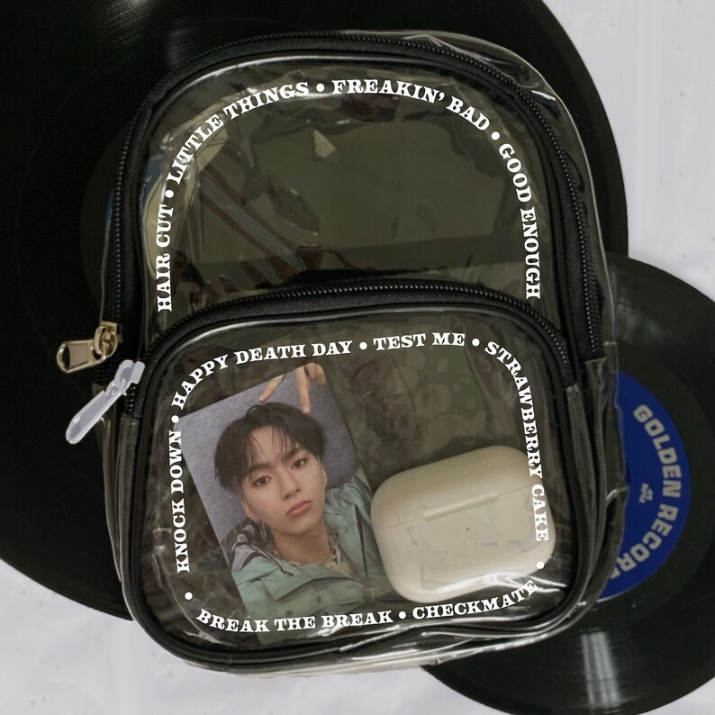 May include: Clear plastic backpack with black trim and white text around the edges. The text reads "Hair Cut Little Things Freakin' Bad Good Enough", "Knock Down Happy Death Day Test Me Strawberry Cake", and "Break The Break Checkmate". The backpack has a photo of a person and a white case inside.