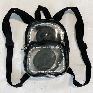 May include: A clear plastic backpack with black straps and white text. The text on the backpack reads: "Cat and Dog, Chasing That Feeling, Oxidovesong, Deja Vu Sugar, Rush Ride, Blue Hour, Drama, Loser Looer, Devil by the Window, Crown."