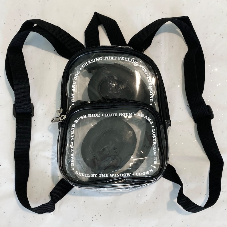 May include: A clear plastic backpack with black straps and white text. The text on the backpack reads: "Cat and Dog, Chasing That Feeling, Oxidovesong, Deja Vu Sugar, Rush Ride, Blue Hour, Drama, Loser Looer, Devil by the Window, Crown."