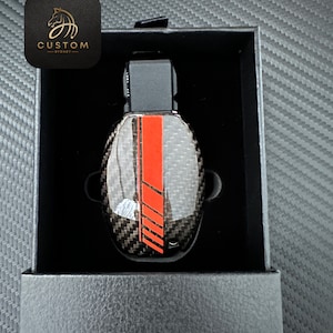 May include: A black and red car key cover with a carbon fiber pattern. The key cover is in a black box with a gold logo that says "CUSTOM - SYDNEY".