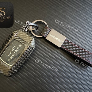 May include: A black carbon fibre key fob with a silver metal insert that reads "LAND CRUISER". The key fob is attached to a black leather strap with red stitching and a silver metal ring.