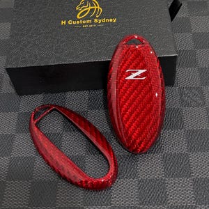 May include: A red carbon fiber key fob cover set featuring a glossy finish and a white Z emblem on the top piece. The set includes a solid top shell and an open bottom frame, presented alongside a black box labeled H Custom Sydney.