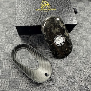 May include: A black carbon fibre key fob with a silver Mercedes-Benz logo and the text "AMG" and "Affalterbach" on it. The key fob is sitting on a black surface.