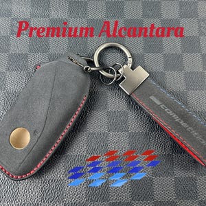 May include: A black leather key fob with red stitching and a silver key ring. The key fob has the word "COMPETITION" printed on it in white lettering. The key fob is attached to a grey and red leather strap with the words "Premium Alcantara" printed on it.