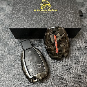 May include: Two black carbon fibre key covers for a Mercedes-Benz car key. One cover has the word "AMG" in red letters. The other cover has a keyhole and four buttons.