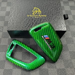 May include: Two green car key covers with a carbon fibre pattern. One cover has the text "M3 Competition" in black and white.